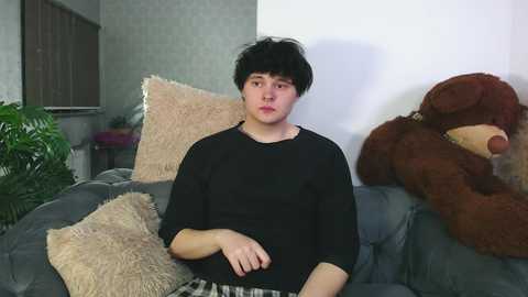 Media: A video of a young person with messy black hair, wearing a black long-sleeve shirt, sitting on a gray couch with plush pillows and a large brown teddy bear. The background shows a gray-patterned wall and a green plant.