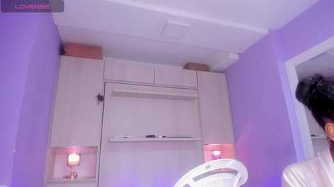 Media: Video of a small, minimalist room with lavender walls, featuring a white TV stand, built-in shelves, and two pink lamps, alongside a white chair.