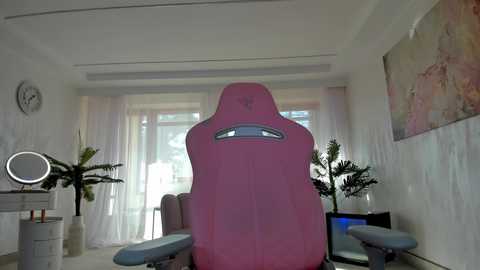 Media: Video of a pink gaming chair with a curved back, in a modern room with white walls, potted plants, and a round mirror.