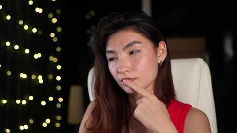 Media: Video of an East Asian woman with long, dark hair, wearing a red dress, contemplating with her finger to her lips in a dimly lit room with a Christmas tree adorned with golden lights in the background.