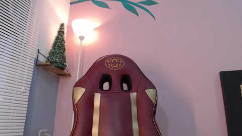 Media: A video of a maroon gaming chair with gold accents, placed against a pink wall with green leaf decals. The chair is positioned next to a small, faux Christmas tree and a lamp.