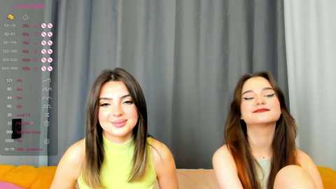 Media: Video of two young women with long brown hair, wearing green tops, sitting on a couch in front of a grey curtain.