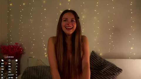 Media: Video of a smiling, topless young woman with long brown hair, sitting on a bed with striped pillows. She has a light complexion and is in a softly lit room with string lights and pink flowers in the background.