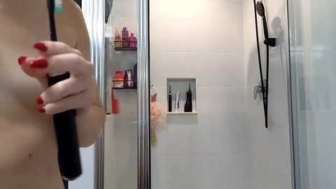 Media: Video of a naked woman with light skin, red-painted nails, holding a black hairbrush in a modern white-tiled bathroom. Shelves hold various beauty products.