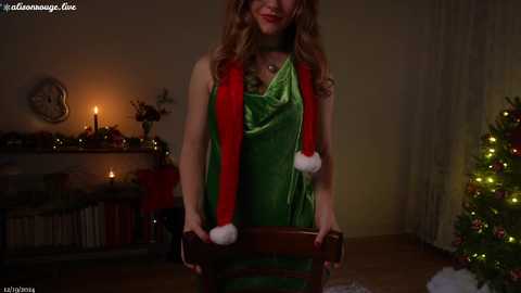 Media: A video of a woman in a green velvet dress with red and white Santa hat, standing in a cozy, warmly lit living room with a lit Christmas tree, candles, and holiday decorations.