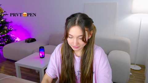 Media: A young woman with long brown pigtails, wearing a lavender t-shirt, sits in a modern living room with a Christmas tree and purple lights, a mobile phone, and a white chair.