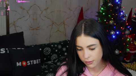 Media: A video of a young woman with long black hair, fair skin, and a pink top, standing in a festive room with a Christmas tree and star-patterned wall.