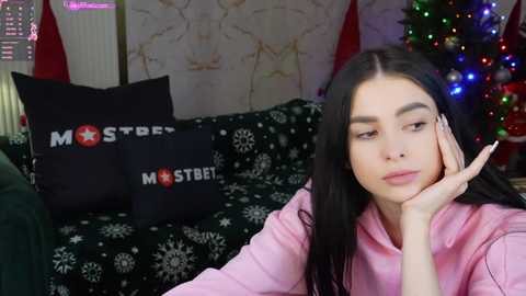 Media: Video of a young woman with long black hair, light skin, and pink hoodie, resting her head on her hand, in a cozy living room with a decorated Christmas tree and black pillows with \"Mister Starbet\" logo.