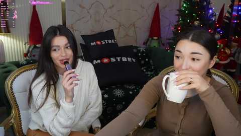 Media: Video of two young women drinking from a cup, one in a white sweater, the other in a brown top, in a cozy, festive living room with a Christmas tree, green couch, and \"Masturbate\" pillows.