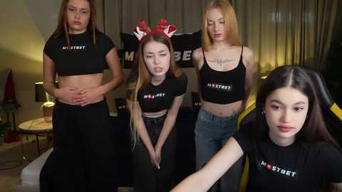 Media: Video of four young women in black \"Mostly\" crop tops, one with antlers, standing in a dimly lit living room, wearing neutral expressions.