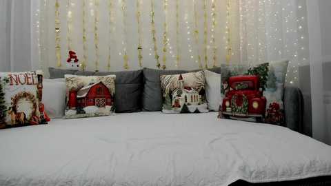 Media: Video of a cozy, festive room with a white bed adorned with holiday pillows featuring rustic cabins and a red truck. A string of gold and white fairy lights hangs on the background, creating a warm, inviting atmosphere.