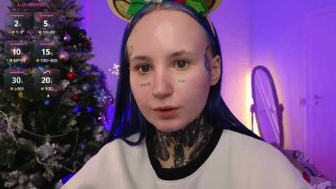 Media: Video of a fair-skinned woman with long blue hair, wearing a green Santa hat and a white hoodie, with detailed tattoo sleeves, in a cozy room with a decorated Christmas tree.