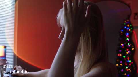 Media: Video of a blonde woman with shoulder-length hair, illuminated by warm lighting, hand in her hair, standing in a room with a Christmas tree and orange wall.