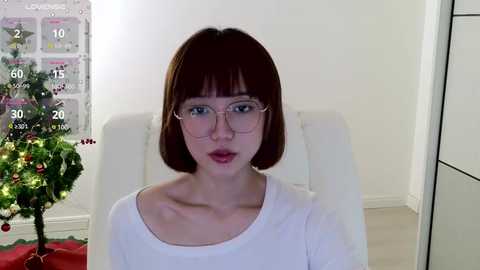 Media: Video of a fair-skinned woman with a bob haircut and glasses, wearing a white sweater, seated in a modern white chair against a plain white wall, with a decorated Christmas tree and calendar visible in the background.