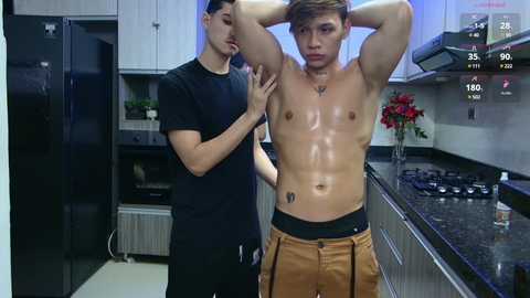 Media: Video of two shirtless Asian men in a modern kitchen. One has a muscular build, the other slender. One massages the other's chest, both wearing only brown shorts. Stainless steel appliances and red flowers in the background.