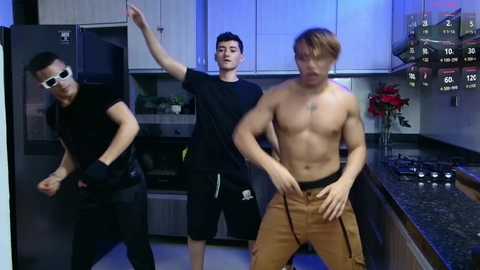 Media: Video of three young men in a modern kitchen: two wear black clothes, one shirtless in tan pants, all dancing.