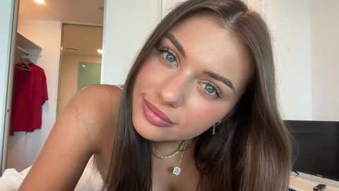 Media: Video of a fair-skinned woman with long brown hair, blue eyes, and a slight smile, wearing a strapless top, in a modern, minimalist bedroom with a closet and a flat-screen TV.