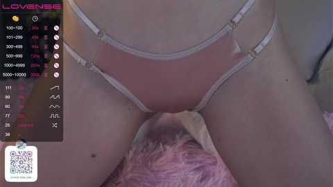 Media: A video of a fair-skinned person wearing sheer white lingerie with strappy details, lying on a pink fur blanket. The background is blurred, with a digital screen displaying a \"Loveress\" profile on the left.