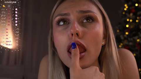 Media: Video of a blonde woman with light skin, green eyes, and blue lipstick, holding her finger to her mouth, in a dimly lit room with a Christmas tree in the background.