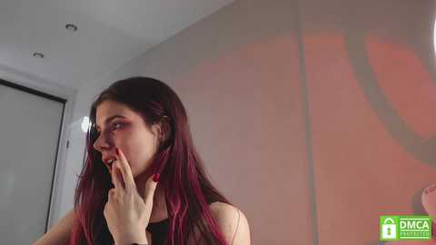 Media: Video of a young woman with long, dark hair, fair skin, and red nails, in a black top, covering her mouth in a bedroom with a beige wall and red lighting.