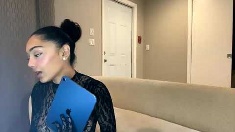 Media: Video of a Latina woman with dark skin and dark hair in a bun, wearing a black lace top, holding a blue tablet and looking pensive against a beige hallway with white door and carpeted floor.