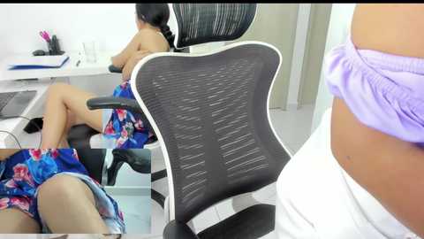 Media: Video of a woman in a white shirt, partially undressed, sitting in a black office chair with mesh backrest, with another woman in a floral dress in the background.