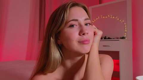 Media: Video of a light-skinned, blonde woman with long hair, resting her chin on her hand, smiling softly. Pink lighting illuminates her in a softly lit room with a vanity mirror and fairy lights in the background.