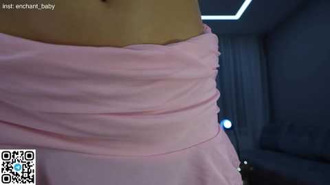 Media: Video of a woman's torso, wearing a light pink, strapless dress with a gathered waist, in a dimly-lit room with modern decor and a QR code in the bottom left corner.