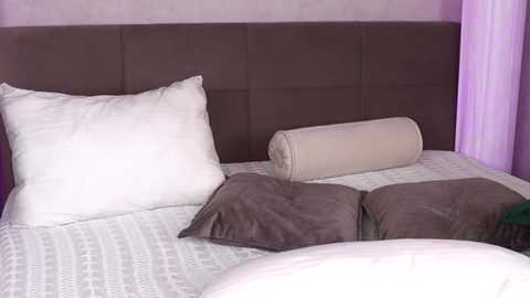 Media: Video of a modern bedroom with a brown upholstered headboard, white pillow, beige rolled-up pillow, gray duvet, white and gray patterned bedspread, and light purple walls.