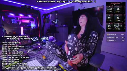 Media: Video of a woman with long black hair in a black and white camo onesie, sitting in a gaming chair with a computer desk cluttered with gaming equipment.