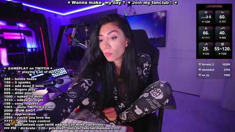 Media: Video of a young woman with long black hair, wearing a black and white patterned top, sitting at a gaming setup with multiple monitors. Text overlays include fan club information and social media handles.