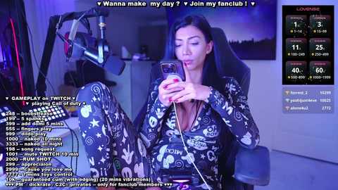 Media: Video of a woman in a black, patterned onesie with a smartphone, seated in a dark room with a Twitch overlay.