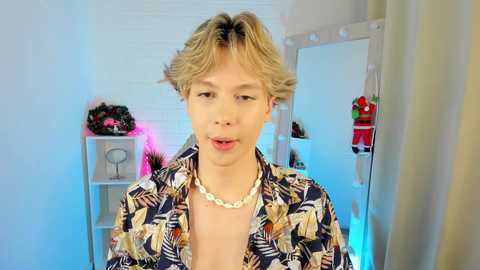 Media: Video of a young, light-skinned male with short, tousled blonde hair, wearing a Hawaiian shirt and shell necklace, standing in a brightly lit room with pink and blue accents, a mirror, and a red dress hanging.