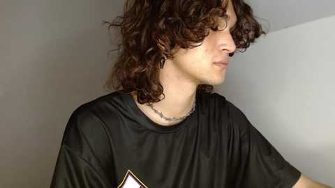 Media: Video of a young man with curly dark hair, fair skin, wearing a black T-shirt and silver chain necklace, standing indoors against a plain white wall, looking slightly to the side.