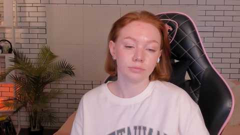 Media: Video of a pale-skinned redhead woman in a white t-shirt, sitting in a black gaming chair with pink accents, against a tiled wall with a potted plant and a lit lamp in the background.