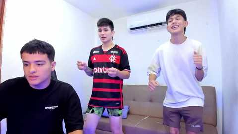 Media: Video of three young Asian men, two wearing soccer jerseys and shorts, one in a white T-shirt, energetically playing a video game in a small, dimly lit room with a beige couch.