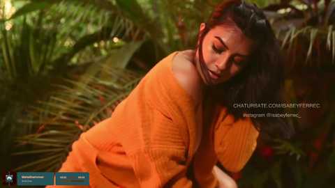 Media: Video of a woman with medium brown skin and dark hair, wearing an orange off-shoulder dress, sitting in lush green foliage.