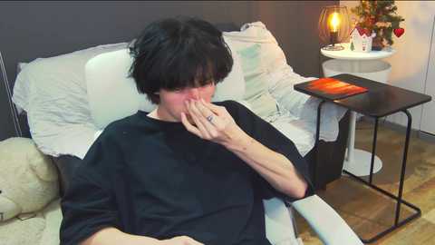 Media: Video of a person with short black hair, wearing a black shirt, covering their face with their hands, sitting on a white chair in a cozy room with a lit table lamp, soft bedding, and a red book.