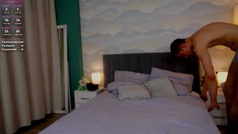 Media: Video of a nude man bending over a bed in a modern bedroom with a gray upholstered headboard and cloud-patterned wallpaper.