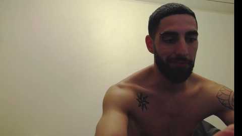 Media: Video of a shirtless, muscular Middle Eastern man with a trimmed beard, dark hair, and tattoos on his shoulders, captured indoors against a plain, light-colored wall.