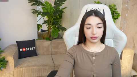 Media: Video of a young woman with long, straight dark hair, wearing a beige long-sleeved top, seated in a white gaming chair, surrounded by green plants and a beige couch with a black pillow reading \"MS STUDY.\