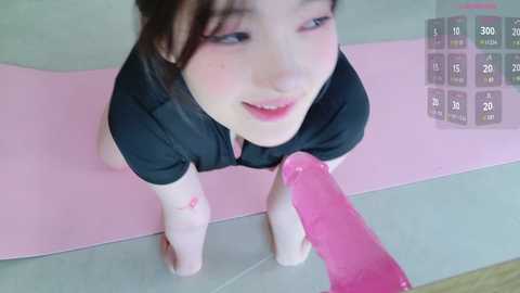 Media: Video of a young Asian woman with fair skin, dark hair, and pink makeup, kneeling on a pink yoga mat, holding a pink dildo, smiling.