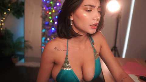 Media: A video of a woman with medium-length dark hair, wearing a turquoise bikini with sequins, leaning over a table. She has a tattoo on her right shoulder. The background features a colorful Christmas tree and a warm light.