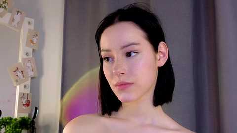 Media: A video of a young East Asian woman with straight black hair, fair skin, and a delicate facial structure, wearing a white top, in a softly lit room with blurred pastel-colored wall art and a plant.