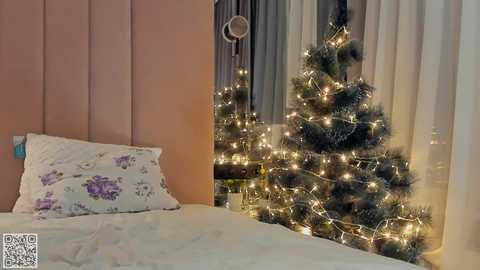 Media: A cozy bedroom with a white bed, floral pillow, and a lit Christmas tree next to a window, adorned with fairy lights. The room has a soft, warm ambiance.