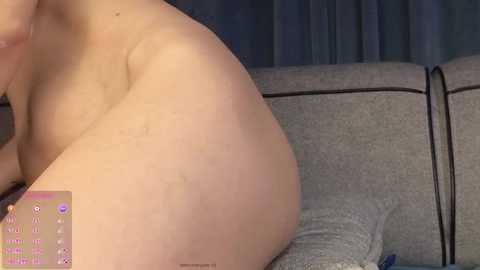 Media: A close-up video of a nude, light-skinned woman's curvaceous buttocks, lying on a grey, textured sofa. Her skin appears smooth with a subtle stretch mark. Dark curtains in the background.