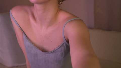 Media: Video of a woman with fair skin and short brown hair, wearing a light gray tank top, leaning forward slightly, showing her collarbone and bare shoulders. The background is blurred, featuring soft, muted tones.