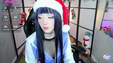 Media: Video of a young woman with long, dark hair in a Santa hat, wearing a black fishnet top and blue jacket, sitting in a gaming chair in a festive room with holiday decor.