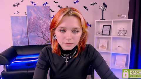 Media: Video of a pale-skinned woman with orange hair, wearing a black top, cross necklace, and piercings, in a Halloween-themed room with bat decorations and white shelves.