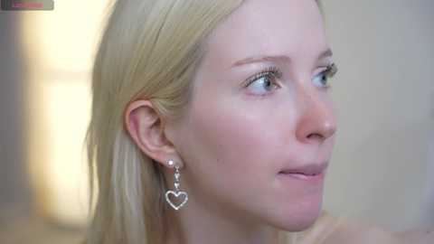 Video of a fair-skinned, blonde woman with blue eyes, wearing a silver heart-shaped earring, looking contemplative against a soft-focus, beige background.